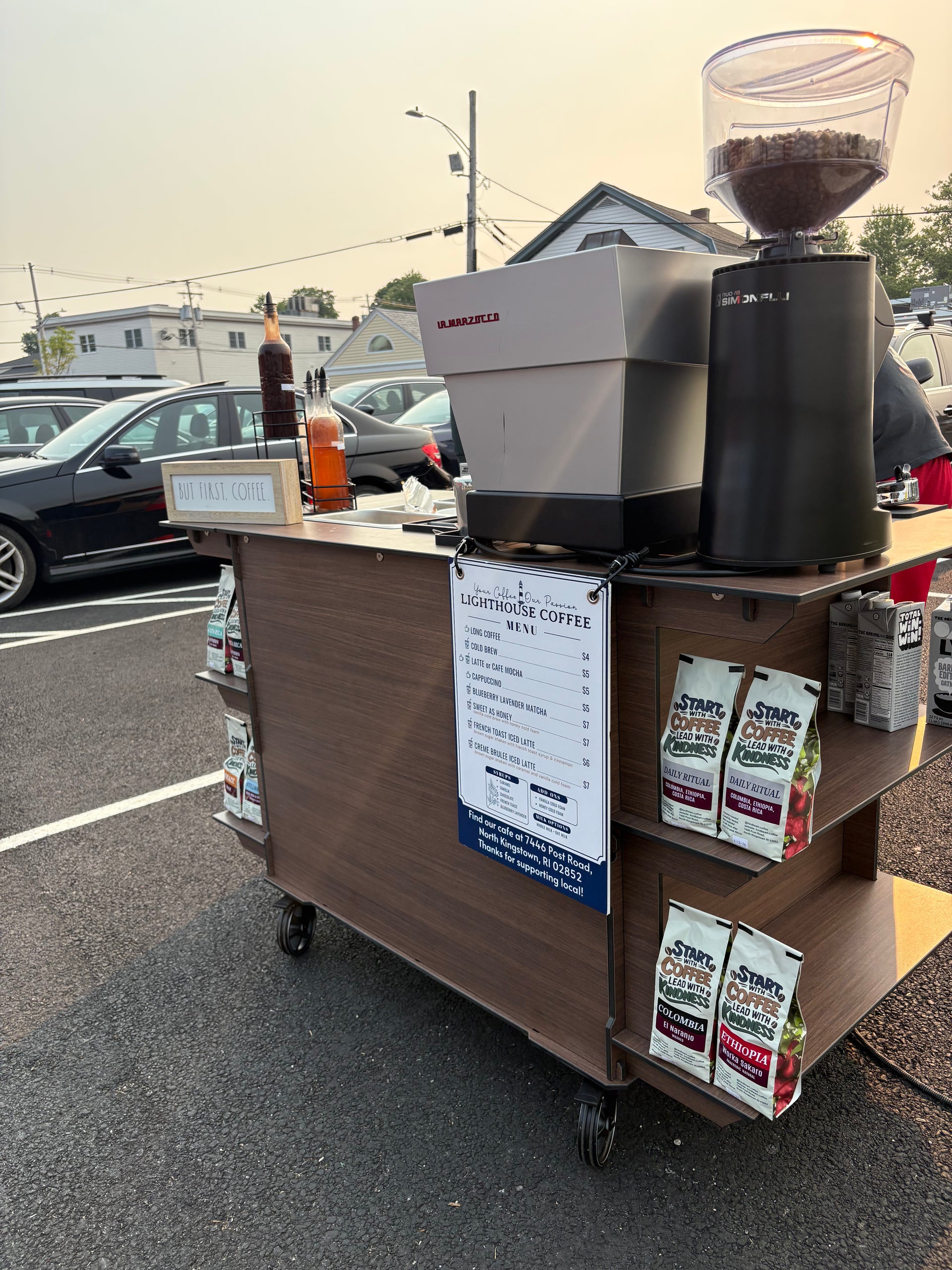 A coffee cart setup outdoors on a parking lot asphalt, featuring a wooden cart with wheels, a white and black espresso machine, a black coffee grinder filled with coffee beans, a menu sign, and bags of coffee on the shelves.