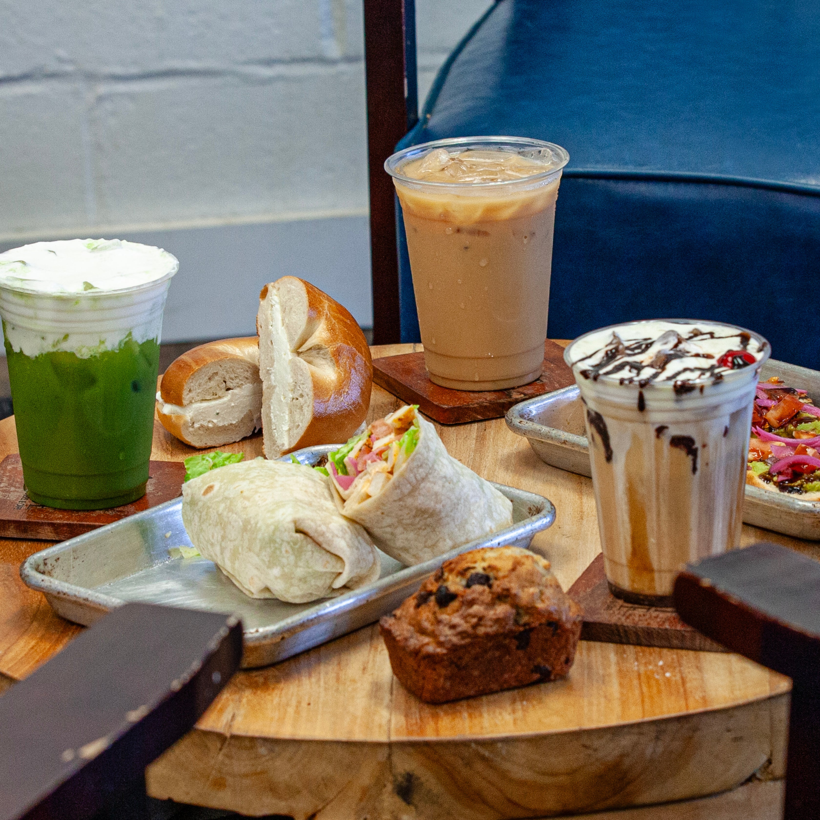 A cafe table setting with multiple food and drink items, including two wraps on a metal tray, a bagel, a muffin, an iced coffee, a green drink with whipped cream, and a chocolate-drizzled milk drink, arranged on wooden coasters and a wood-grain table.