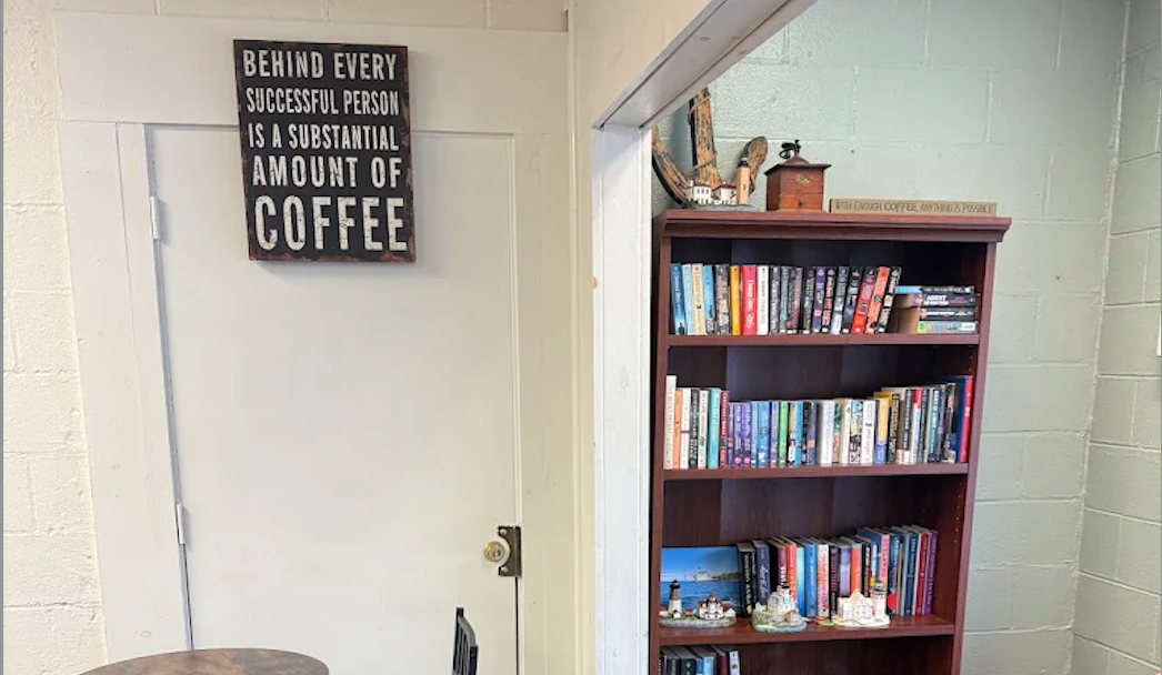 A dark brown wooden bookcase filled with books stands against a white brick wall next to a white door with a decorative sign that reads 'BEHIND EVERY SUCCESSFUL PERSON IS A SUBSTANTIAL AMOUNT OF COFFEE'.